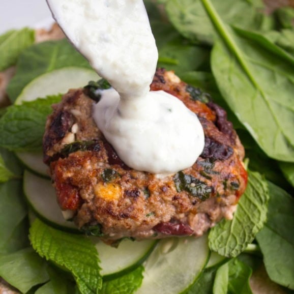 Greek sun-dried tomato lamb burger patty with yogurt sauce on top, sitting on a bed of cucumber and spinach