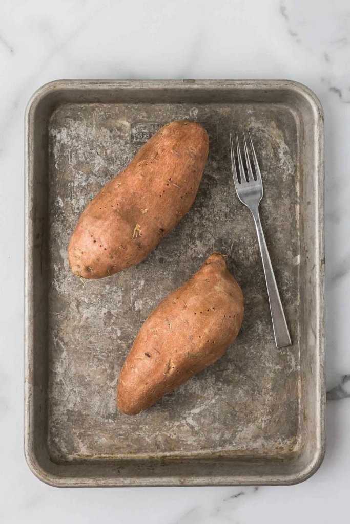 two sweet potatoes on a metal sheet pan with a fork