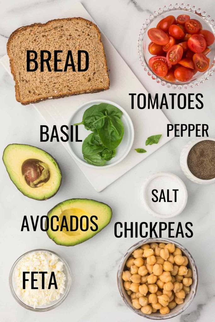 bread, chickpeas, feta, avocados, basil, and tomatoes on a white marble countertop