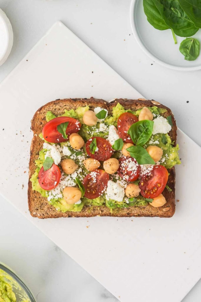 a slice of avocado toast with feta cheese, chickpeas, and cherry tomatoes on parchment paper