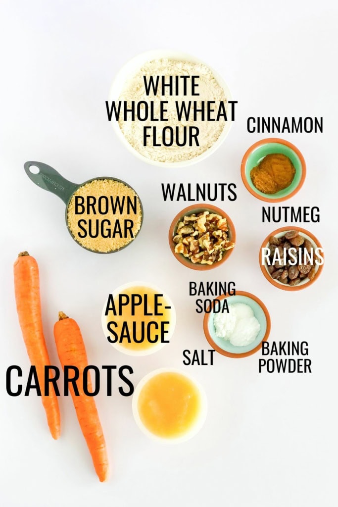 carrots, applesauce, white whole wheat flour, brown sugar, walnuts, raisins, and more baking ingredients on a white surface