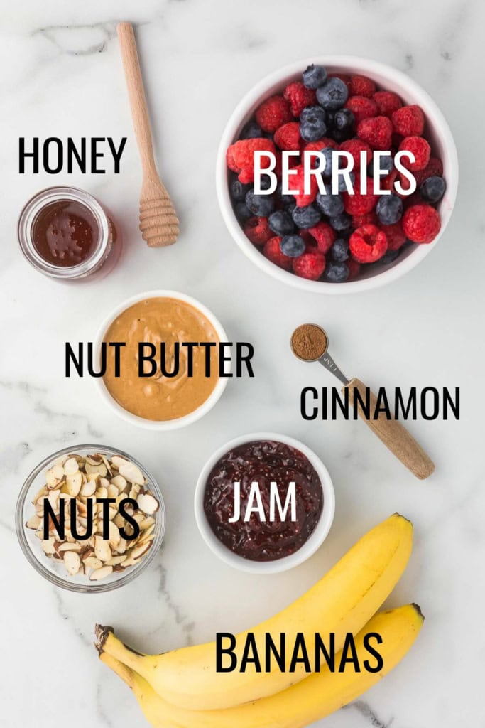 sweet toppings: berries, honey, jam, bananas, nuts, nut butter, and cinnamon in small bowls on a white marble surface