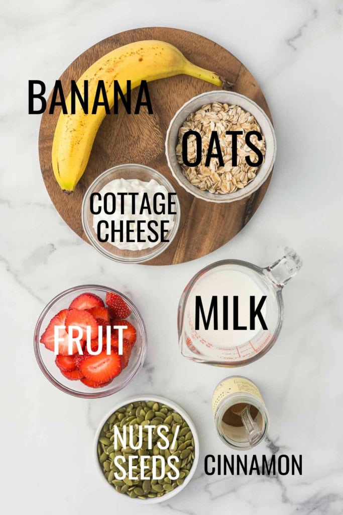 banana, oats, cottage cheese, milk, cinnamon, sliced strawberries, and pumpkin seeds in small bowls on a white marble background